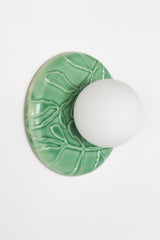 Jade green ceramic globe sconce close-up, glossy leaf and branch relief backplate with frosted white globe offset, modern chinoiserie botanical wall light