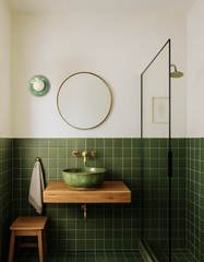 Minimal modern bathroom with deep olive-green square tile walls and a floating light-wood vanity topped with a green ceramic vessel sink, brass wall-mount faucet, and round brass-framed mirror. The Jade Green Lierre ceramic wall sconce by Sazerac Stitches adds a sculptural botanical accent on the white upper wall, featuring a glossy leaf-relief backplate and a soft white globe shade. A black-framed glass shower screen, warm brass shower head, and a simple wooden stool create a clean spa-like look with Scand