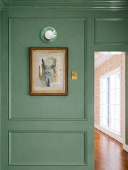 Jade green Lierre ceramic wall sconce with raised leafy relief pattern and frosted globe shade, mounted on a deep green paneled wall with classic picture frame molding and brass switch plate, styled with vintage framed artwork and warm wood floors, handcrafted small business lighting designed and assembled in New Orleans with ceramic made in North Carolina, traditional classic cottage English style hallway or living room wall sconce