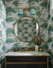 See the handmade double Marcella Sconce styled in a jewel-box powder room — its two fluted, hand-glazed ceramic shades in a soft pastel green celadon-style glaze glowing against dramatic crane-and-pine wallpaper. The scalloped, Deco-inspired shades feel unabashedly feminine, and the celadon-style glaze pools deeper in the flutes while catching light on the raised ribs, reading as a delicate green that turns almost aqua in the light. Paired with warm brass, green marble, and moody black cabinetry, it's a les