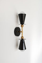 Side angle of the Janus Sconce in matte black with brass hardware, a double headed two light wall sconce with stacked cone shades and clean disc details, shown installed vertically with globe bulbs, a sculptural architectural statement sconce for modern bathrooms, bedside wall lighting, hallway lighting, and contemporary mid century modern interiors that want bold graphic lighting