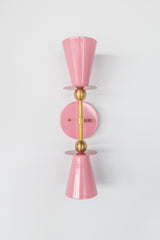 Front view of the Janus Sconce in blush pink with brass arm and accents, featuring two opposing tapered cone shades with crisp disc plates and soft globe bulbs, a graphic geometric modern wall sconce inspired by mid century modern and brutalist design, ideal for flanking a mirror, lighting a powder room or bathroom vanity, modern entryway lighting, and minimalist or color pop interiors