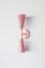Blush pink Janus Sconce, a bold architectural two light wall sconce with stacked tapered cone shades, flat disc details, and warm brass hardware, shown mounted vertically with globe bulbs, a modern sculptural brutalist meets mid century modern wall light for bathroom vanity lighting, hallway sconces, bedroom bedside lighting, and statement lighting in design forward interiors