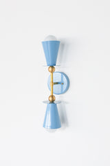 The Janus Sconce is a geometric, double-coned wall light in celestial blue with brass hardware. Two matching cone shades point away from each other — one up, one down — each sitting on a flat brass disc above a brass ball joint. The cone shape gives it an Art Deco feel, while the exposed round bulbs and brass ball stem lean mid-century modern. A round backplate anchors it all to the wall, with the whole thing reading clean, symmetrical, and graphic.