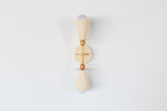 Studio photo of the Janus Sconce in cream and brass, a playful two-light wall sconce with opposing cone shades, white globe bulbs, a round backplate, and sculptural brass ball accents. The geometric shape feels part mid century modern, part art object, making it a fun choice for maximalist interiors, dopamine decor, eclectic powder rooms, colorful bedrooms, kids spaces, hallways, and quirky designer lighting schemes. Shown in cream with brass, available in all Sazerac Stitches powder-coated colors.