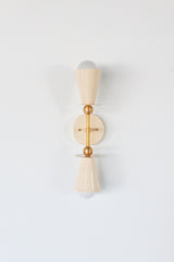 Straight-on product photo of the Janus wall sconce in cream with brass hardware, featuring two opposite-facing cone shades, white globe bulbs, and a balanced vertical silhouette. The fixture feels graphic, quirky, and architectural, with enough softness to work in a pretty powder room and enough structure to fit mid century modern or eclectic interiors. Use it as a statement wall sconce in a bathroom, bedroom, hallway, reading nook, kids room, or color-drenched maximalist space.