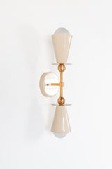 Angled studio view of the Janus Sconce showing the depth of the cream powder-coated cone shades, brass stem, round wall plate, and playful stacked brass sphere details. This sculptural wall light adds personality without feeling overly fussy, with a geometric silhouette that works in modern bathrooms, bedside lighting, creative hallways, art-filled interiors, maximalist rooms, and dopamine decor spaces. A customizable colorful sconce for anyone who wants lighting that feels designed, not basic.