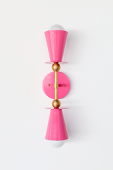 This vertical bath bar sconce is built to flank a bathroom mirror in pairs, with two Doll Pink cone shades and brass ball hardware giving it a mid-century, Art Deco-adjacent shape. It works as vanity lighting in a powder room, a girl's bathroom or nursery, or as hallway sconces. Bold color, symmetrical shape, made to actually get noticed instead of disappearing into the wall.