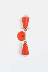 Angled view of flame orange Janus wall sconce with brass mount and globe bulb, sculptural modern lighting with double cone shade silhouette, statement bathroom vanity sconce, bedside reading light, or hallway accent for bold color drenched interiors.