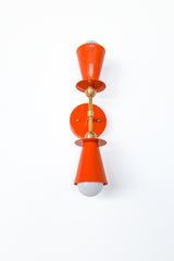 Angled view of flame orange Janus wall sconce with brass mount and globe bulb, sculptural modern lighting with double cone shade silhouette, statement bathroom vanity sconce, bedside reading light, or hallway accent for bold color drenched interiors.