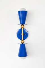 The Janus Sconce in Mediterranean Blue takes a classic double-cone silhouette and saturates it in deep cobalt, with brass ball joints holding the two shades apart from a round backplate. One cone points up, one points down, so the exposed bulbs bookend the fixture at either end. It reads confident enough to anchor a powder room or hallway on its own, no extra styling required.