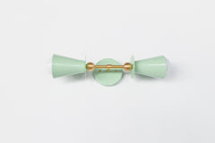 Angled view of the Janus Sconce in glossy yellow with brass backplate and center rod, showing stacked geometric double cone shades with soft globe bulbs, a sculptural modern wall sconce with mid century modern and brutalist design influence, ideal as a bathroom wall sconce, mirror side lighting, hallway statement light, bedroom wall light, or designer accent fixture in color forward interiors.