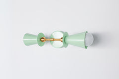 Angled view of the Janus Sconce in glossy yellow with brass backplate and center rod, showing stacked geometric double cone shades with soft globe bulbs, a sculptural modern wall sconce with mid century modern and brutalist design influence, ideal as a bathroom wall sconce, mirror side lighting, hallway statement light, bedroom wall light, or designer accent fixture in color forward interiors.