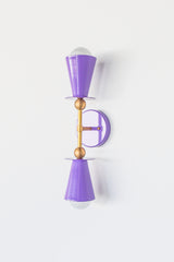 Janus wall sconce with glossy orchid purple powder-coated metal shades, warm brass hardware, and white glass globes shown from an angled side view against a white background.  Orchid purple wall sconce with warm brass accents and white glass globes. Sculptural modern lighting inspired by Art Deco styling for colorful interiors.