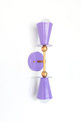 Three-quarter view of the Janus wall sconce featuring glossy orchid purple powder-coated metal shades, warm brass hardware, white glass globes, and a sculptural modern profile against a white background. Sculptural orchid purple wall sconce with brass accents and white glass globes. Modern statement lighting for bathrooms, bedrooms, hallways, and colorful interiors.