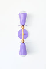 Front view of the Janus wall sconce with glossy orchid purple powder-coated metal shades, warm brass hardware, white glass globes, and a matching round backplate on a white background.  Modern orchid purple wall sconce with brass accents and a sculptural silhouette. Perfect for colorful bathrooms, bedrooms, hallways, and eclectic interiors.
