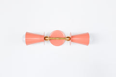 Studio photo of the Janus Sconce in peach and brass, a playful two-light wall sconce with opposing cone shades, white globe bulbs, a round backplate, and sculptural brass ball accents. The geometric shape feels part mid century modern, part art object, making it a fun choice for maximalist interiors, dopamine decor, eclectic powder rooms, colorful bedrooms, kids spaces, hallways, and quirky designer lighting schemes. Shown in peach with brass, available in all Sazerac Stitches powder-coated colors.