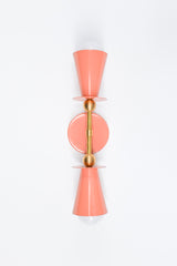 Studio photo of the Janus Sconce in peach and brass, a playful two-light wall sconce with opposing cone shades, white globe bulbs, a round backplate, and sculptural brass ball accents. The geometric shape feels part mid century modern, part art object, making it a fun choice for maximalist interiors, dopamine decor, eclectic powder rooms, colorful bedrooms, kids spaces, hallways, and quirky designer lighting schemes. Shown in peach with brass, available in all Sazerac Stitches powder-coated colors.
