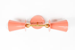 Studio photo of the Janus Sconce in peach and brass, a playful two-light wall sconce with opposing cone shades, white globe bulbs, a round backplate, and sculptural brass ball accents. The geometric shape feels part mid century modern, part art object, making it a fun choice for maximalist interiors, dopamine decor, eclectic powder rooms, colorful bedrooms, kids spaces, hallways, and quirky designer lighting schemes. Shown in peach with brass, available in all Sazerac Stitches powder-coated colors.