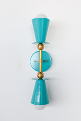Symmetry is the whole point of the Janus Sconce: two matching teal cone shades point in opposite directions from a central brass stem, each one finished with its own brass ball joint. The round teal backplate keeps the hardware simple so the color and shape do the work. It reads equally at home in a playful powder room or as a graphic moment in a hallway.