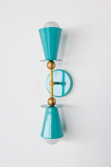 The Janus Sconce takes a classic double-cone silhouette and gives it a colorful update in teal powder coat, with warm brass ball joints holding the two shades apart from a round backplate. One cone faces up, one faces down, so the exposed bulbs read almost like punctuation at either end. It's the kind of fixture that can carry a bathroom, hallway, or bedroom on its own, no extra decorating required.