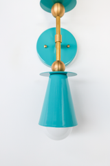 A closer look at what makes the Janus Sconce feel considered rather than generic: the brass ball joints on either side of the round teal backplate, the visible mounting screws, and the glossy powder-coat finish on the cone shade itself. Small details like this are usually what separate a fixture that photographs well from one that actually holds up in person.