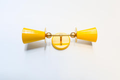 Yellow & Brass Janus Sconce, a bold architectural two light wall sconce with stacked tapered cone shades, flat disc details, and warm brass hardware, shown mounted vertically with globe bulbs, a modern sculptural brutalist meets mid century modern wall light for bathroom vanity lighting, hallway sconces, bedroom bedside lighting, and statement lighting in design forward interiors