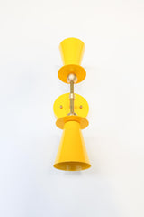 Yellow & Brass Janus Sconce, a bold architectural two light wall sconce with stacked tapered cone shades, flat disc details, and warm brass hardware, shown mounted vertically with globe bulbs, a modern sculptural brutalist meets mid century modern wall light for bathroom vanity lighting, hallway sconces, bedroom bedside lighting, and statement lighting in design forward interiors