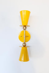 Yellow & Brass Janus Sconce, a bold architectural two light wall sconce with stacked tapered cone shades, flat disc details, and warm brass hardware, shown mounted vertically with globe bulbs, a modern sculptural brutalist meets mid century modern wall light for bathroom vanity lighting, hallway sconces, bedroom bedside lighting, and statement lighting in design forward interiors