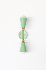 Front view of the Janus Sconce in Vista Green and brass, a two light wall sconce with stacked cone shades, disc details, and globe bulbs in a bold architectural silhouette, ideal as a mid century modern bathroom vanity light, modern hallway wall light, bedroom wall sconce, or sculptural statement lighting for colorful, design forward interiors.