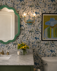 This bright, playful bathroom showcases the Adele Two Light Sconce in Poolside and brass, mounted on whimsical animal wallpaper in soft blue, green, and neutral tones. A wavy green mirror, marble vanity top, brass plumbing fixtures, yellow flowers, and a colorful framed painting give the room a fresh, lively feel that is perfect for a kid-friendly bathroom or colorful powder room. The sconce adds a sculptural touch while tying together the room’s blue and green palette, making it a great fit for eclectic de