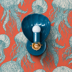 A deep lagoon-blue shell sconce against a coral-orange jellyfish wallpaper — complementary color contrast doing all the heavy lifting. The Coquille from Sazerac Stitches is a glossy scallop-shell wall light with a warm brass fitting and scalloped floral backplate, in a rich teal-blue set off by a whimsical vintage-inspired jellyfish print on warm coral. Playful and a little theatrical, made for a maximalist bathroom, an eclectic powder room, or a bedroom with personality. Works for blue and orange color sch