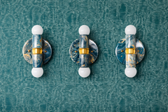 Three Thalia sconces in lagoon, cream, and gold marbled finish displayed side by side on teal textured wallpaper. Each compact two-light sconce has a glossy marbled round backplate, a matching marbled cylinder, a brass center band, and white globe bulbs at the top and bottom. A bold, art-object lighting moment that works in powder rooms, hallways, entryways, or modern eclectic and coastal-inspired interiors where color and pattern are the main event.