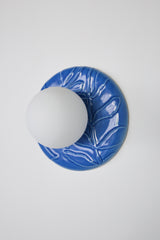 Lierre ceramic wall light in Sky Blue celadon-style glaze, round ivy leaf relief plate with matte white globe, shown illuminated on wall with soft ambient halo, hardwired decorative sconce lighting