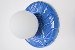 Lierre ceramic wall light in Sky Blue celadon-style glaze, round ivy leaf relief plate with matte white globe, shown illuminated on wall with soft ambient halo, hardwired decorative sconce lighting