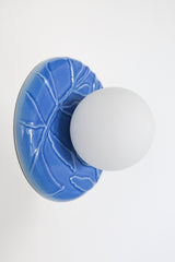 Lierre Sconce installed as a ceramic wall sconce in Sky Blue celadon-style glaze, raised ivy leaf relief plate with matte white 4 inch globe diffuser, shown lit with warm glow on a textured wall