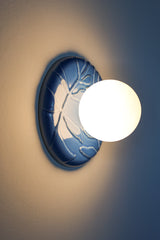 Lierre ceramic wall light in Sky Blue celadon-style glaze, round ivy leaf relief plate with matte white globe, shown illuminated on wall with soft ambient halo, hardwired decorative sconce lighting