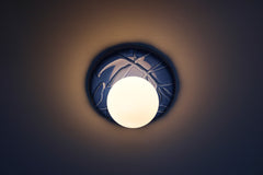 Lierre ceramic wall light in Sky Blue celadon-style glaze, round ivy leaf relief plate with matte white globe, shown illuminated on wall with soft ambient halo, hardwired decorative sconce lighting