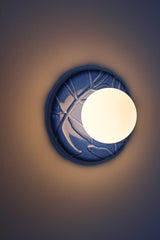 Lierre ceramic wall light in Sky Blue celadon-style glaze, round ivy leaf relief plate with matte white globe, shown illuminated on wall with soft ambient halo, hardwired decorative sconce lighting