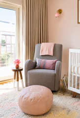 Soft, light-filled nursery reading corner featuring a blush pink Loa sconce with warm brass hardware mounted on a pale pink wall above a gray glider chair. Neutral floor-length drapery, a textured cream rug, blush leather pouf, white crib, and a small round wood side table with pink flowers create a cozy baby girl nursery with modern, feminine, and softly boho style. This pastel nursery design highlights a blush pink wall sconce, colorful nursery lighting, and a stylish reading nook, making it ideal for mod