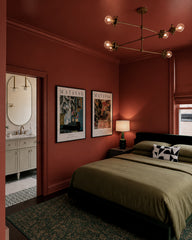 Moody color-drenched bedroom with rust and terracotta walls and ceiling, styled like a boutique hotel. A brass Linear Octavia Chandelier hangs overhead with exposed globe bulbs for warm, sculptural modern bedroom lighting. The space features olive green bedding, framed Matisse art prints, and a window with a tailored Roman shade. An open doorway reveals a bright bathroom with an arched mirror and brass accents, creating a high-contrast mix of modern vintage, mid-century modern, and art deco inspired decor.