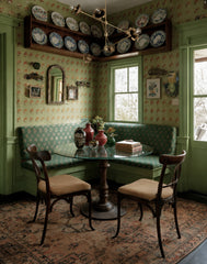 Vintage-inspired green breakfast nook with patterned wallpaper, built-in banquette seating, antique rug, and round glass dining table beneath a brass multi-arm modern chandelier. The eclectic corner dining space features mismatched wooden chairs, decorative wall plates, and colorful vases for a cozy, maximalist interior design look.