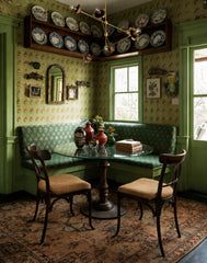 Vintage-inspired green breakfast nook with patterned wallpaper, built-in banquette seating, antique rug, and round glass dining table beneath a brass multi-arm modern chandelier. The eclectic corner dining space features mismatched wooden chairs, decorative wall plates, and colorful vases for a cozy, maximalist interior design look.
