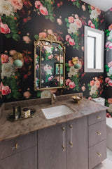 Feminine ultra glam bathroom with dark floral wallpaper, an antique style gold mirror, brass and lucite drawer pulls and handles, and mint and brass globe sconces