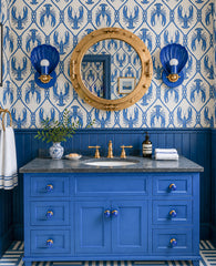 Playful coastal bathroom with blue lobster wallpaper, saturated blue wainscoting, a matching blue vanity, brass porthole mirror, gray marble countertop, and striped blue floor tile. The Mediterranean Blue Coquille sconces bring shell-shaped bathroom lighting into the room, tying the nautical wallpaper and brass fixtures together. This image is strong for coastal bathroom lighting, lobster wallpaper bathroom, blue vanity ideas, shell wall sconces, nautical powder room, grandmillennial bathroom decor, and col