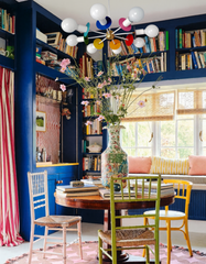 Eclectic dining nook with the Luz chandelier featuring playful multicolor accents and globe bulbs, hanging above a round wooden table surrounded by mismatched painted chairs. The vibrant space includes built-in navy blue bookcases filled with books, a patterned pink rug, striped red curtains, and a large decorative vase centerpiece, blending bold statement lighting with maximalist interior design.