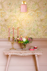 The Marcella sconce in glossy pastel pink ceramic with a brass backplate glows softly against a vintage-inspired floral and bird wallpaper, illuminating a romantic vignette featuring a pale pink-painted wall, an ornate weathered console table, a gold candelabra with pink taper candles, a lush floral arrangement in a glass vase, and delicate tea accessories—perfect for eclectic, feminine interiors or maximalist cottagecore styling.