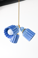 A small chandelier with fluted, scalloped-edge cone shades in a deep periwinkle blue, connected by smooth glass balls in a softer sky blue, all on slim brass hardware. The two-tone blue palette and delicate fluted shape make this a strong pick for a nursery, a breakfast nook, or a powder room that wants something more collected than a standard glass pendant. Shown here at an angle with one bulb lit, giving a sense of how the shades splay outward from the central brass fitting.