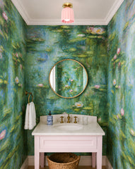 A light pink ceramic Marcella flushmount with brass canopy brings soft, sculptural color to a tiny powder room wrapped in dreamy water lily wallpaper. The space pairs a round brass mirror and brass faucet with a pale marble countertop and a blush pink vanity, creating a pretty, elevated mix of pastel color and warm metal. Great inspiration for anyone searching for a pink ceramic flush mount ceiling light, brass bathroom ceiling fixture, powder room lighting, or a colorful, wallpaper-forward, grandmillennial