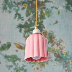 A blushed-pink ceramic shade cascades downward in elegant vertical pleats, crowned with a white sphere finial and brass gallery band. The glaze catches light in a luminous, almost boudoir-like glow. Against botanical wallpaper with hints of garden green and golden accents, this blush reads romantic and intimate—a fixture designed for someone who believes in color without apology, but prefers whisper to shout.
The soft pink has a mature, collected quality—not precious, but intentional.